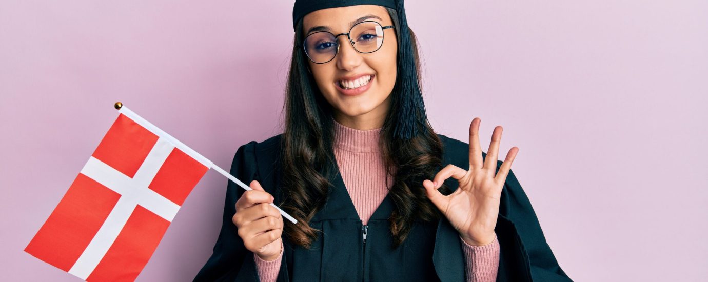 Young hispanic woman wearing graduation uniform holding denmark flag doing ok sign with fingers, smiling friendly gesturing excellent symbol